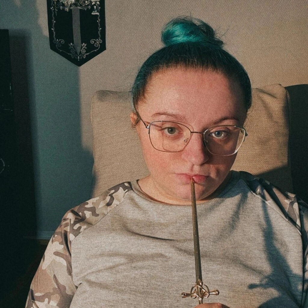 White girl with a green bun. Wearing glasses and a camo type of blouse. Closer to her mouth she holds a letter opener in the form of a small sword.