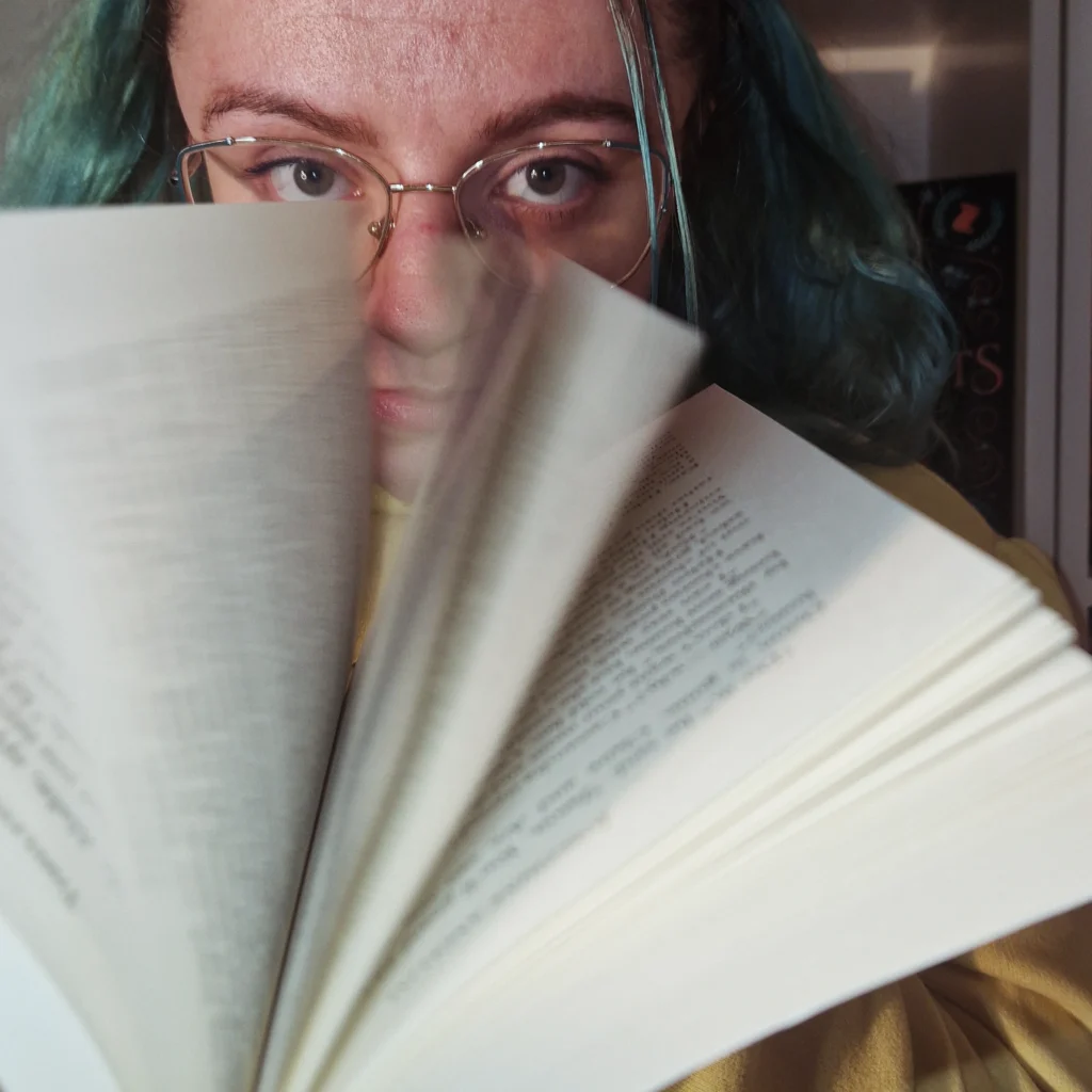 White woman with green hair, and glasses is holding a book. While taking the picture she is flipping through it and the caption is a book with the pages flipped.